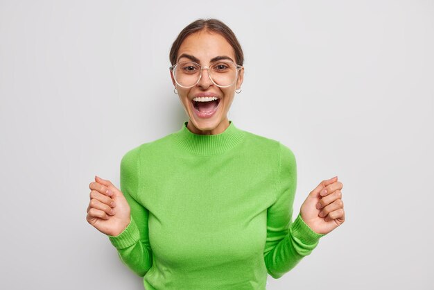 Positive brunette woman raises hands exclaims loudly wears transparent eyeglasses and casual green jumper feels very happy hears positive news isolated over white background Emotions concept