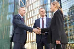 Free photo positive business colleagues meeting in city, standing outdoors and shaking hands near office building. low angle shot. agreement and partnership concept