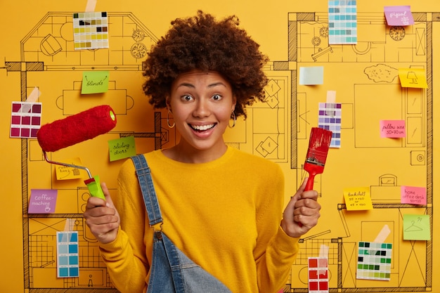 Free Photo positive busy female painter holds paint roller and brush, does house repairing, dressed in yellow jumper and overalls