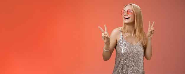 Free photo positive charming european carefree stylish girlfriend having fun dancefloor show victory peace signs look up happily smile in sunglasses glamour glittering dress enjoy celebration red background