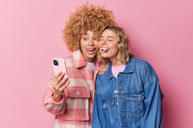 Free photo positive cheerful women make grimace at camera of smartphone stick out tongue foolish around take selfie dressed in fashionable clothes isolated over pink background make photo of themselves