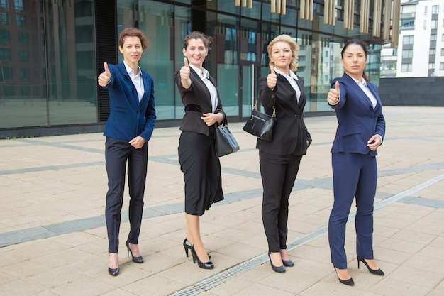 Free Photo positive confident businesswomen team standing together near office building, showing like gesture, making thumb up, looking at camera. full length. teamwork and business success concept