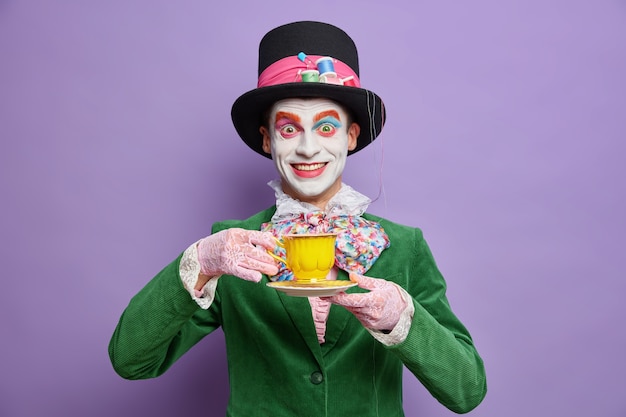 Free Photo positive crazy hatter wears bright colorful makeup enjoys drinking tea on party dressed in costume celebrates halloween poses happy against purple wall