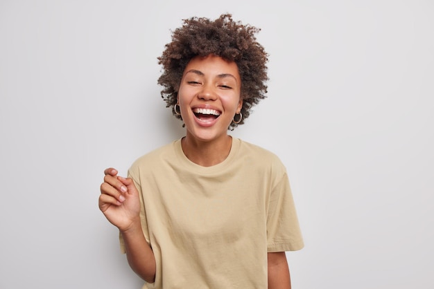 Positive curly haired beautiful woman laughs happily has carefree expression keeps mouth opened wears casual beige t shirt isolated over white studio background Sincere human emotions concept