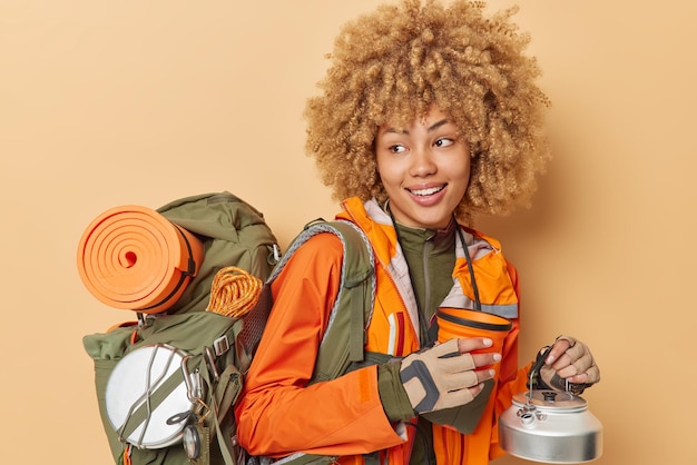 Free photo positive curly haired female tourist poses with camping utensils looks gladfully away dressed in windbreaker carries rucksack full of necessary equipment isolated over beige background hiking concept