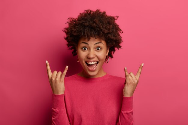 Positive curly haired girl makes horn gesture, enjoys rock n roll music, laughs with joy, wears casual jumper