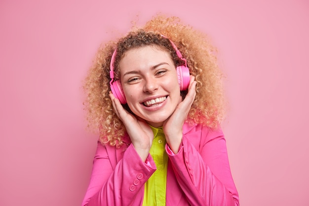 Free Photo positive curly haired teenage girl enjoys listening favorite music wears stereo headphones being in good mood wears bright clothes isolated over pink wall. happy female meloman listens song