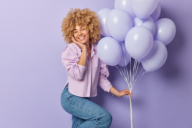 Positive curly haired woman celebrates special occasion feels very happy gets congratulations holds bunch of inflated balloons wears shirt and jeans isolated over purple background Party time