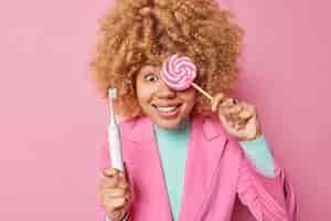 Free photo positive curly haired woman covers eye with delicious candy holds electric toothbrush has sweet tooth dressed in elegant clothes isolated over pink background teeth problem and harmful food