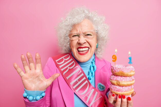 Positive curly haired woman pensioner raises palm feels very happy holds pile of glazed doughnuts celebrates 91st birthday wears festive clothes