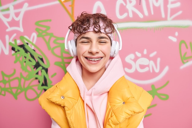 Positive curly haired youngster smiles gladfully has braces on teeth dressed in hoodie with vest enjoys favorite music via wireless headphones draws graffiti on street walls without permission