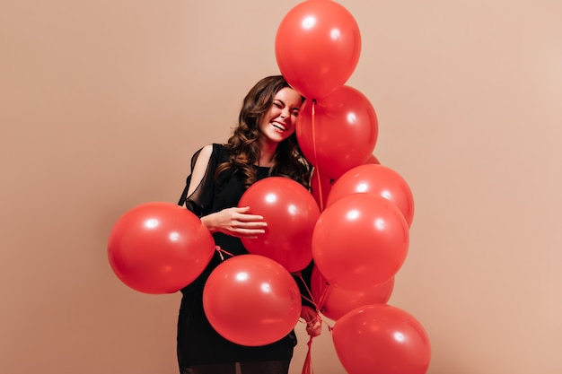 Free photo positive curly woman in black outfit laughs and poses with red balloons on isolated background.