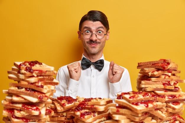 Free Photo positive customer rejoices all inclusive in restaurant, stands near pile of delicious toasts, wears elegant white shirt and bowtie, clenches fists, 