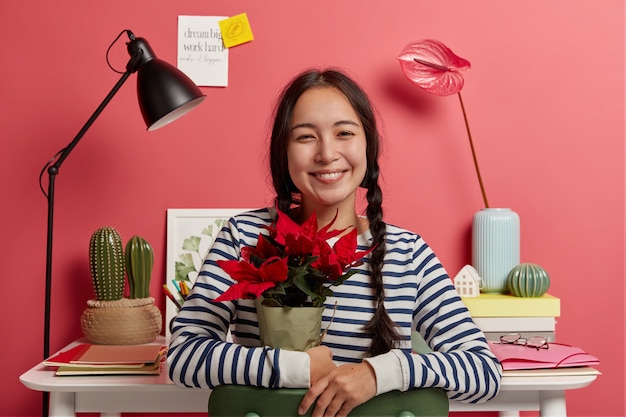 Positive dark haired girl holds red potted flower, poses against cozy coworking space