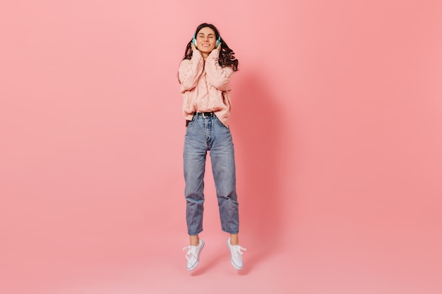 Positive dark-haired girl smiling while listening to music in blue headphones. Woman in knitted outfit and white sneakers jumping on pink background.