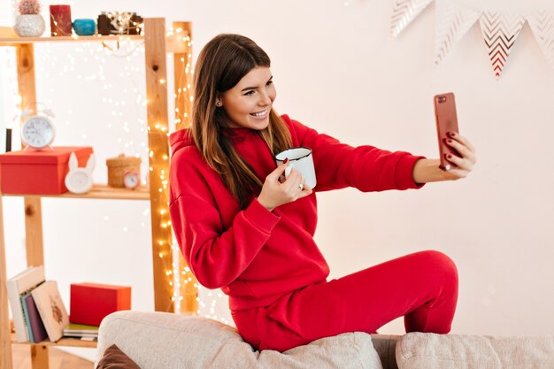 Positive dark-haired woman taking selfie. Studio shot of attractive young lady drinking tea.