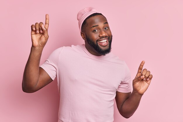 Positive dark skinned man with thick beard dances carefree raises arms has good mood