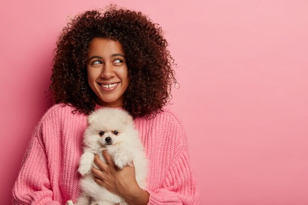 Positive dark skinned teenage girl with Afro bushy hairstyle, poses with white spitz in pink studio, thinks about having picnic in nature.