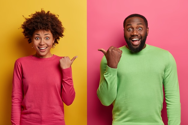 Free photo positive dark skinned woman and man point at each other with thumbs, wear basic clothes, have good mood, suggest picking friend, isolated on yellow and pink wall. look at my companion.