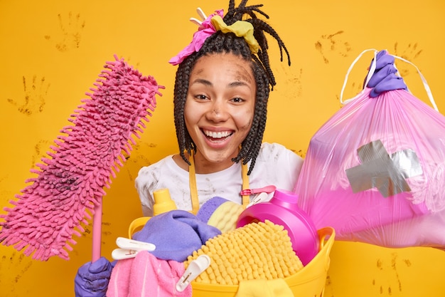 Positive dark skinned woman stands dirty after overall cleaning does housework holds garbage bag and dirty mop tidies up apartment washes laundry smiles broadly isolated over yellow wall