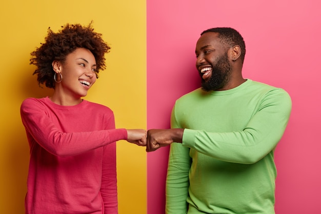 Positive dark skinned young woman and man bump fists, agree to be one team, look happily at each other, celebrates completed task, wear pink and green clothes, pose indoor, have successful deal