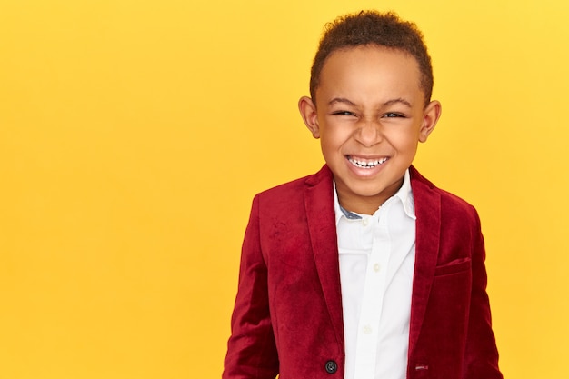 Positive emotions, joy and happy childhood concept. Handsome cheerful dark skinned male child in crimson velvet jacket being in good mood, looking at camera with radiant smile, showing white teeth