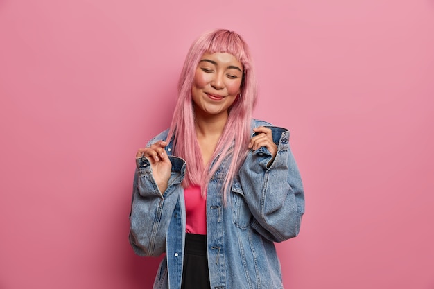 Free photo positive emotions, people and fashion concept. smiling glad young woman with pink long hair, closes eyes, happy to buy new denim jacket, stands indoor