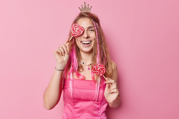 Free photo positive european woman covers eye with heart shaped lollipop wears festive dress and small crown on head smiles broadly has white perfect teeth poses against pink background time for party