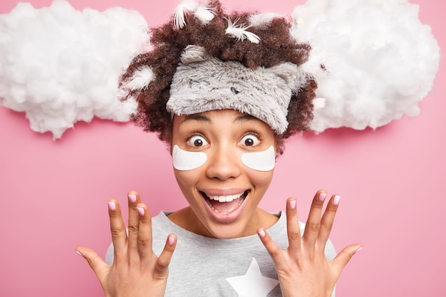 Positive excited curly haired woman raises hands smiles happily has amazed expression feathers stuck in hair wears sleepmask nightwear being surprised by great news isolated over pink wall