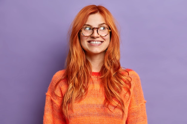 Positive facial expressions. Happy smiling woman with natural red hair looks away and smiles broadly wears spectacles orange jumper.