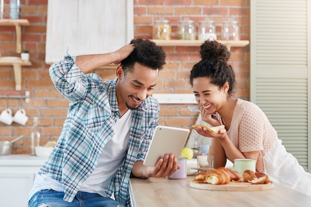 Positive family couple smiles broadly as watches comedy on tablet computer, use free internet connection at home,