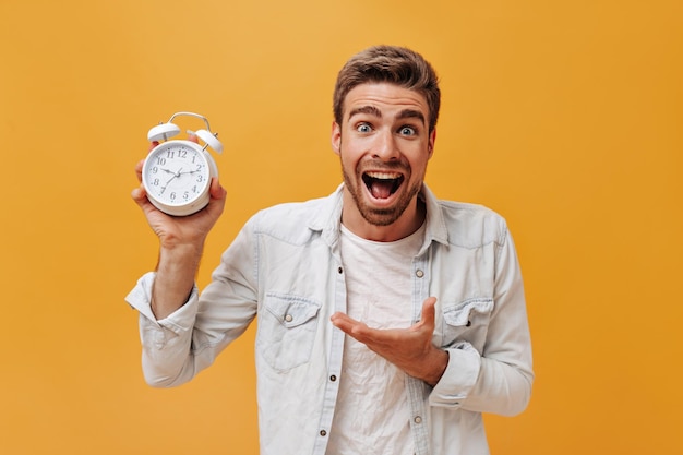 Free photo positive fashionable guy with brown hair and red beard in white tshirt and denim modern jacket showing at big clock and looking into camera