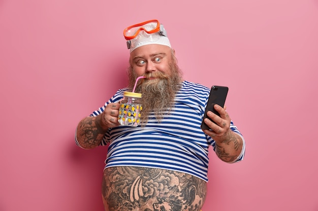 Free Photo positive fatso guy drinks fresh water, takes photo via cellphone, wears swimming snorkeling mask, undersized sailor shirt