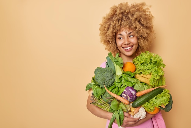 Free photo positive female farmer carries harvested vegetablesbeing agriculture store owner looks away with happy expression isolated over beige background empty space for your promotion healthy food concept