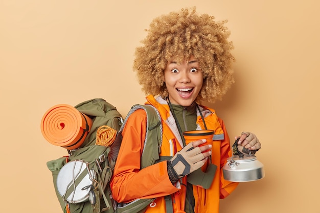 Free photo positive female hiker poses with utensils laughs gladfully dressed in orange jacket carries rucksack leads active lifestyle being in good mood isolated over beige background has vacation in mountains