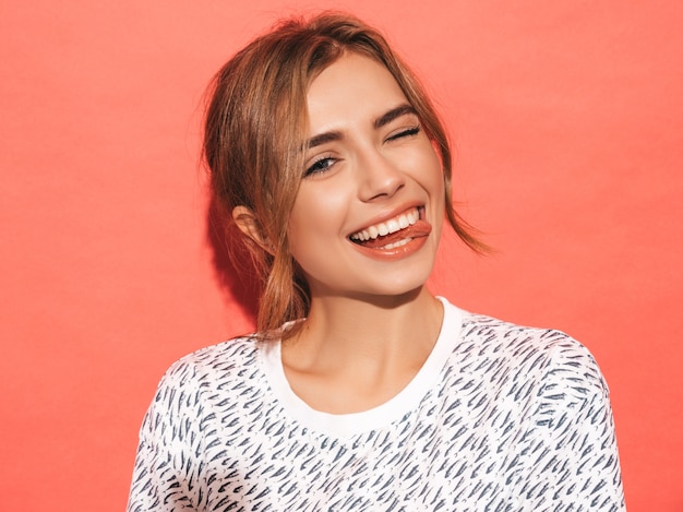 Positive female smiling. Funny model posing near pink wall in studio.Shows tongue and winks