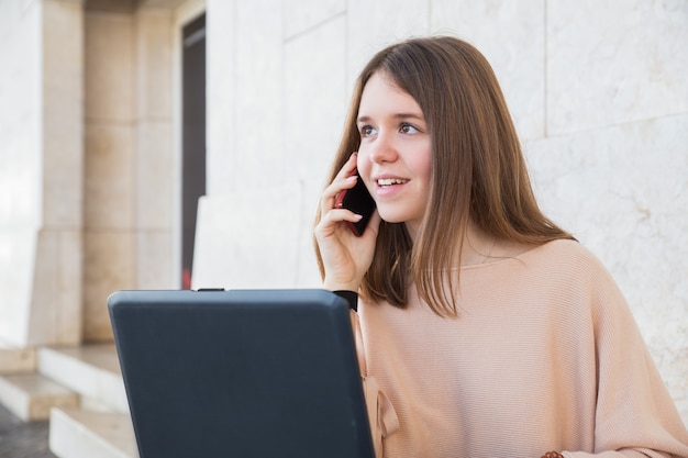 Free photo positive female teenager using laptop and phone at building wall