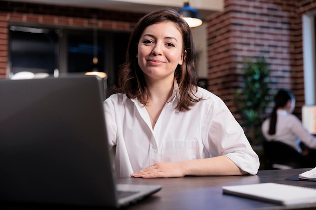 Positive friendly business company executive manager sitting inside agency space. Caucasian young adult woman sitting in office workspace while smiling heartily at camera.