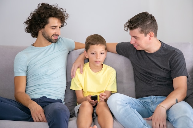 Positive gay dads and son watching TV show together at home, sitting on couch in living room, smiling and hugging boy. Family and home entertainment concept