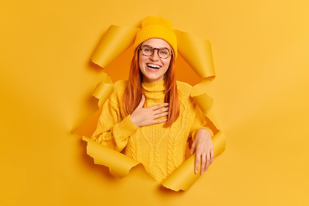 Free Photo positive ginger female model expresses sincere emotions keeps hand on chest feels gratitude smiles broadly wears yellow clothes breaks through paper ripped hole.