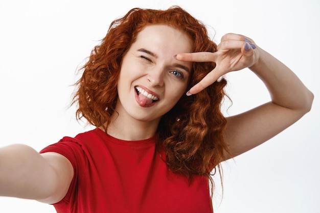 Positive ginger girl with curly hairstyle showing tongue and vsign over eye taking selfie or record video on smartphone front facing camera standing against white background