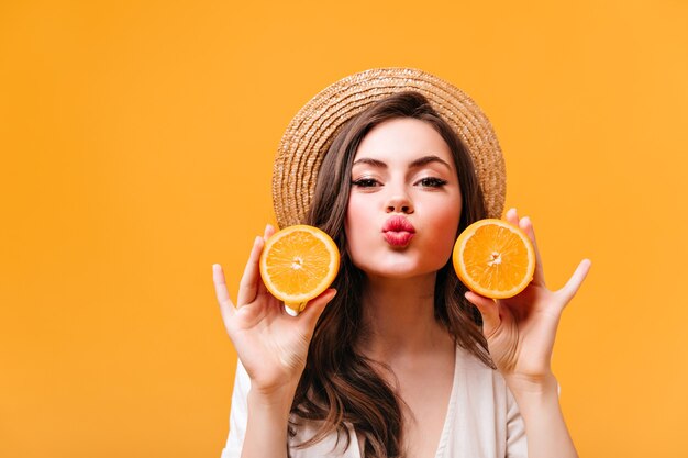 Positive girl in straw hat blows kiss, looks into camera and holds halves of orange.