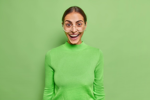 Free photo positive glad european female model feels surprised giggles happily wears transparent glasses and turtleneck pses against vivid green wall