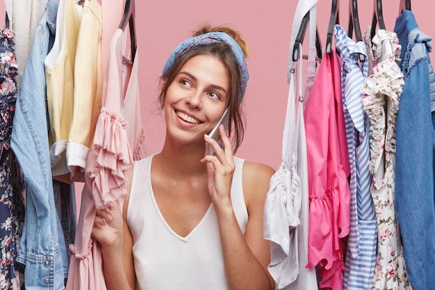 Positive glad woman with pleasant smile, having conversation over smart phone while standing between hangers with clothes. Happy female dressed casually smiling while talking over mobile phone