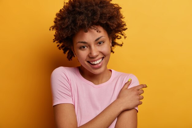 Positive good looking curly haired woman with curly hair, smiles pleasantly, expresses good emotions, has charming look, dressed in casual outfit, poses over yellow wall. Happiness concept