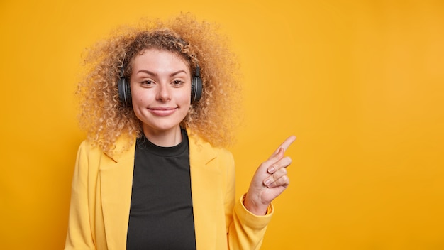 Free photo positive good looking curly haired young woman in formal clothes points away on blank space gives recommendation suggests to place your advertisement here uses wireless headphones poses indoor