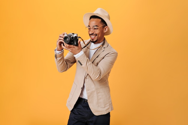 Positive guy in glasses and hat is holding retro camera and smiling on orange wall