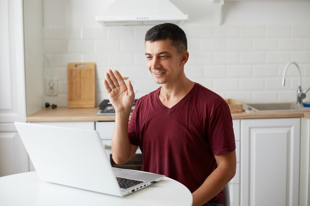 Positive handsome man wearing casual style attire sitting at table in kitchen in front of laptop, having video call, waving hand to web camera, saying hello or good bye.