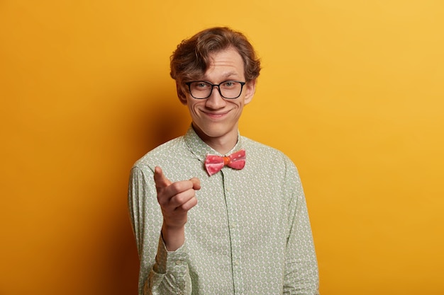 Positive handsome smiling man indicates at you, points with index finger directly , wears fashionable shirt, selects someone, wears glasses, poses over yellow wall. You should join me