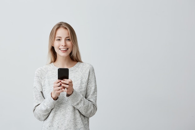 Positive happy blonde-haired woman with broad smile using cell phone, glad to receive message with good news, checking newsfeed on her social network accounts. Modern technologies and communication